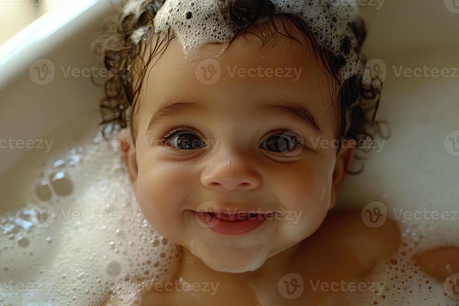 A happy baby with curly hair taking a bath with bubbles and looking at the camera smiling sweetly photo