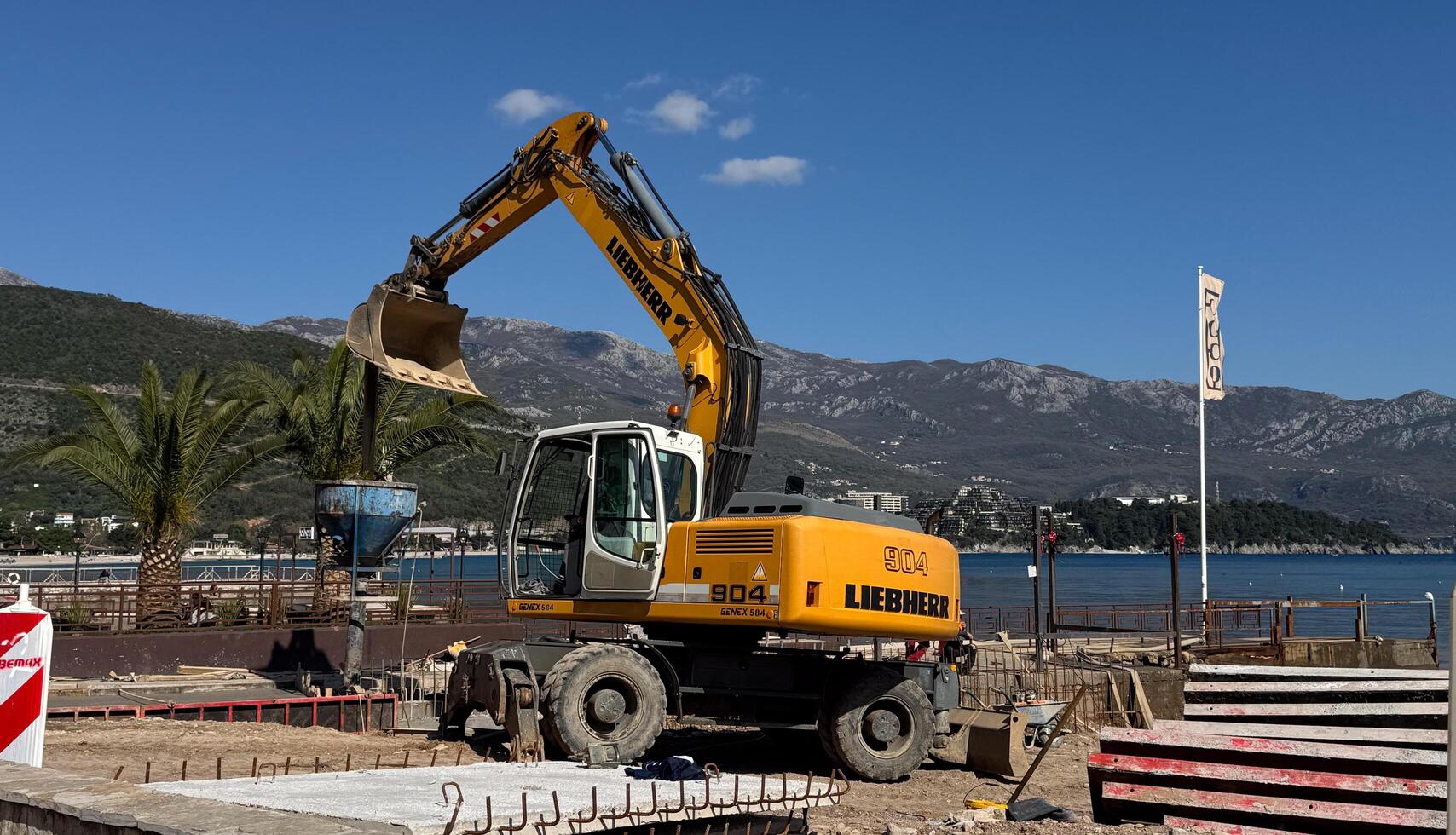 Budva,Montenegro-April 11,2025.A yellow Liebherr excavator working near the coast filling a concrete crater at a coastal construction site with palm trees and a COCO cafe flag. High quality photo