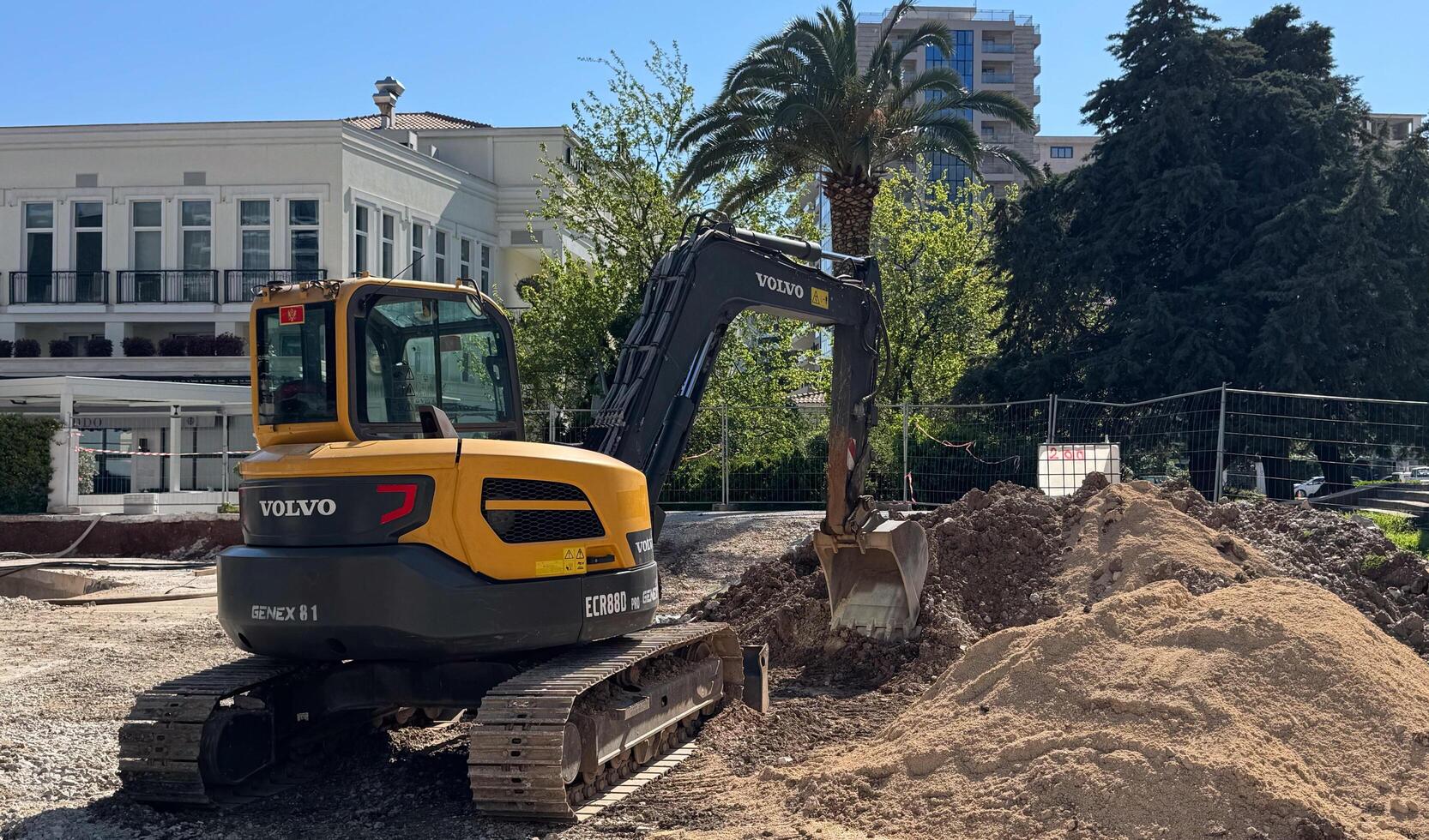 Budva,Montenegro-April 11,2025. Yellow and black Volvo excavator digging soil at a city construction site surrounded by buildings and palm trees. photo