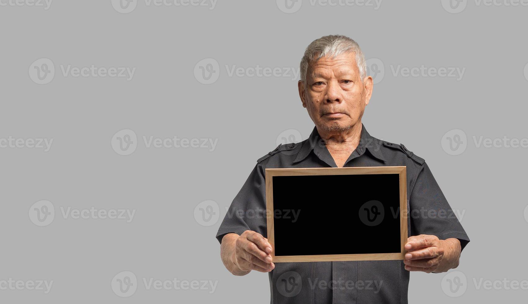 Portrait of an elderly Asian man holding a mini blackboard and looking at the camera with a smile while standing on a gray background. photo