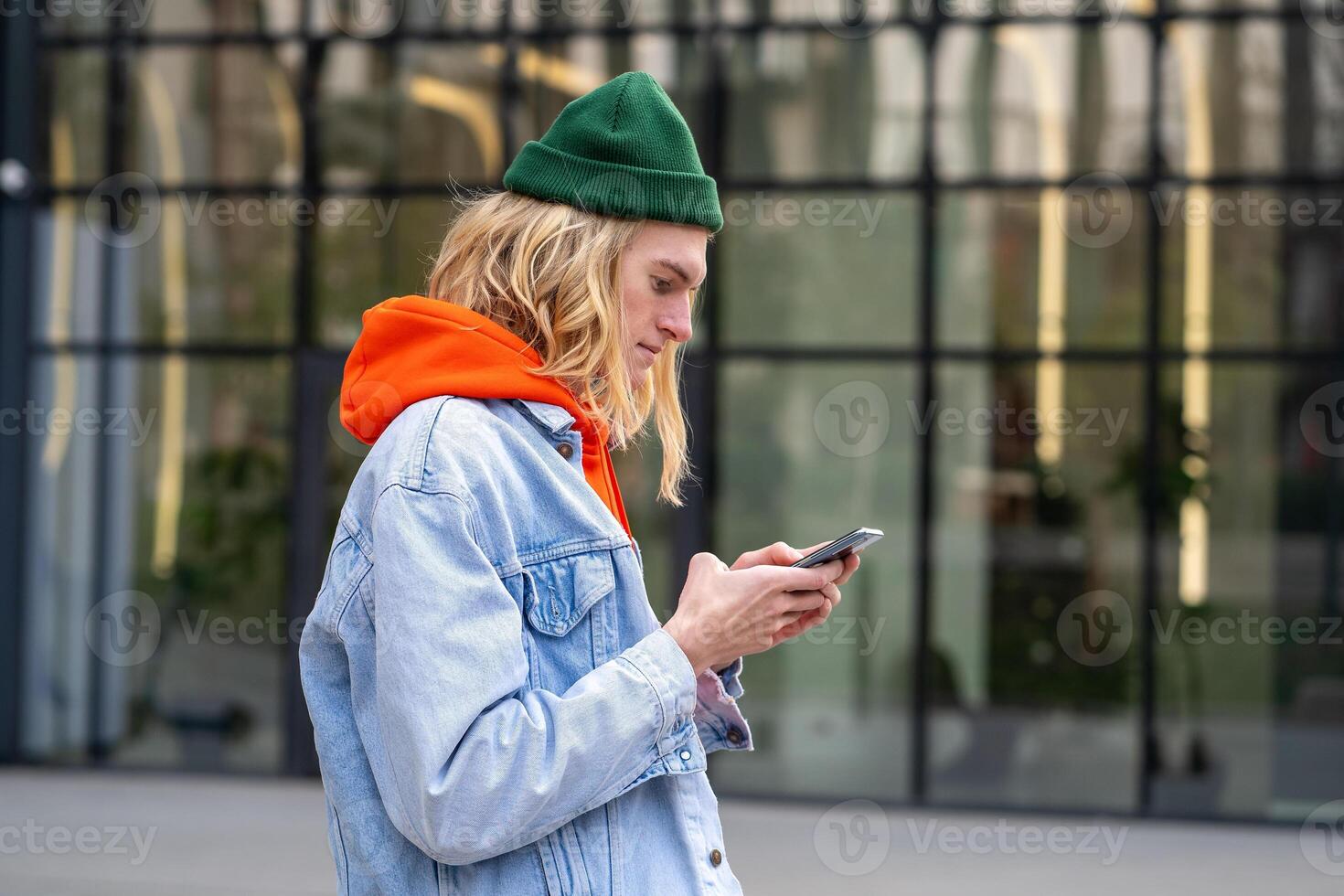 Pleased hipster guy standing outside with smartphone, looking screen typing message in social media. photo