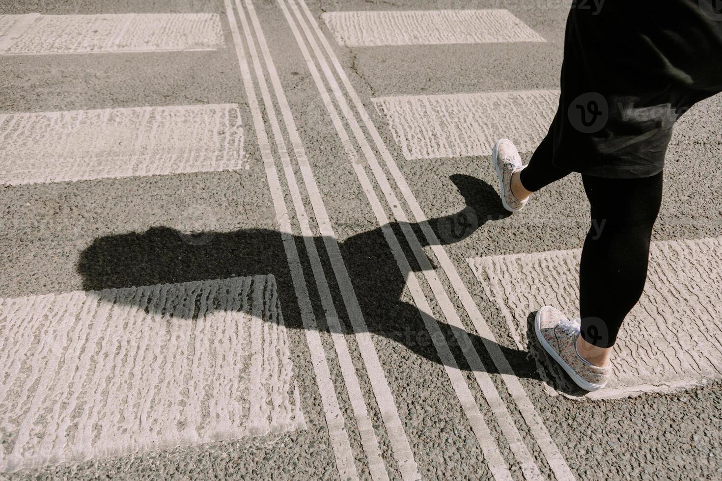 Woman walking across a pedestrian crosswalk with a clear shadow cast on the pavement. Perfect for themes of city life, independence, movement, and everyday moments in urban environments. photo