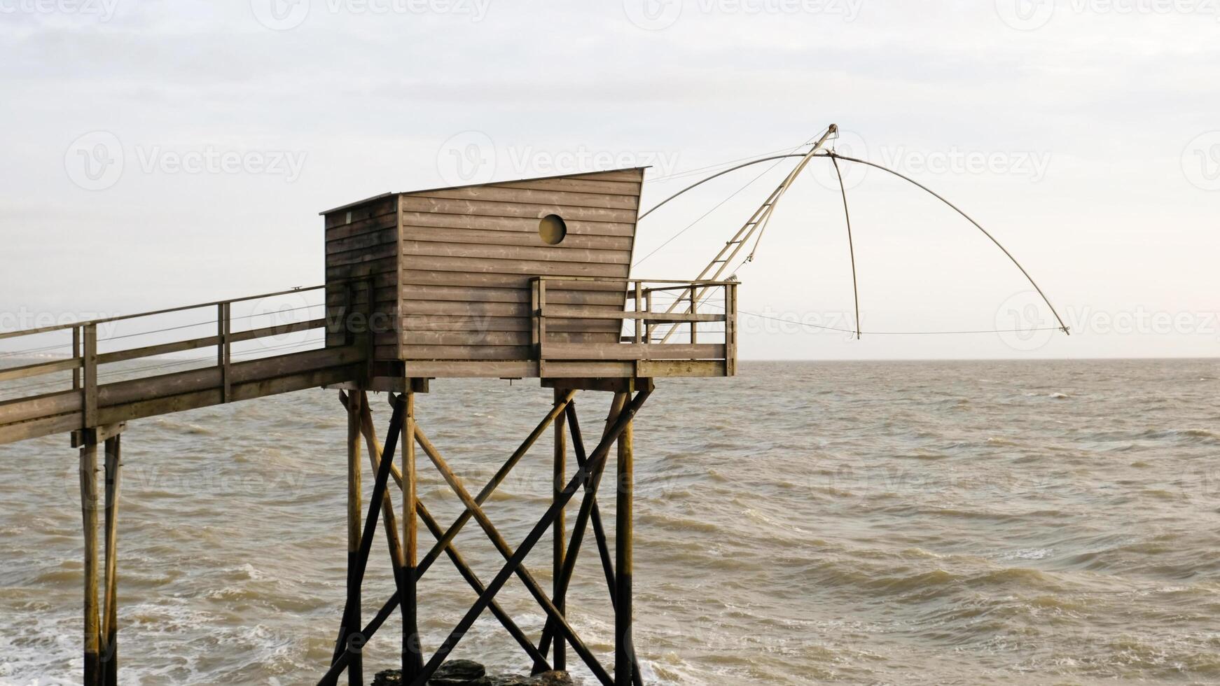 Traditional French fishing hut on stilts with carrelet net over ocean. Wooden structure stands on rocky shore, with waves and cloudy sky. Ideal for projects to travel, close-up photo