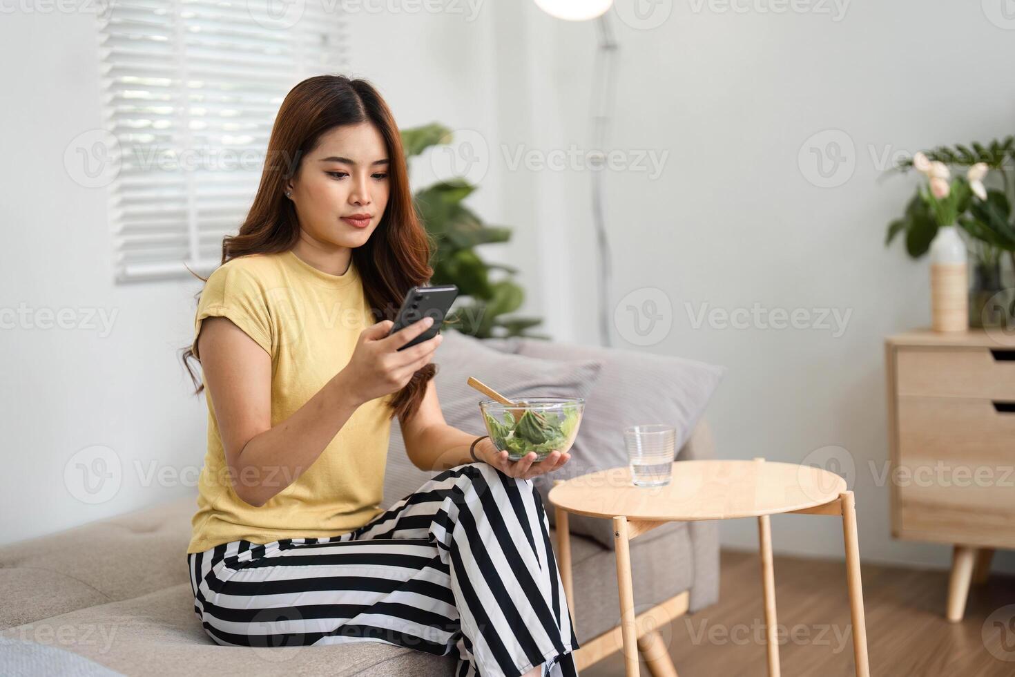 Morning Routine and Healthy Habits. A young woman enjoying a salad while using her phone, embracing healthy living in a cozy setting. photo