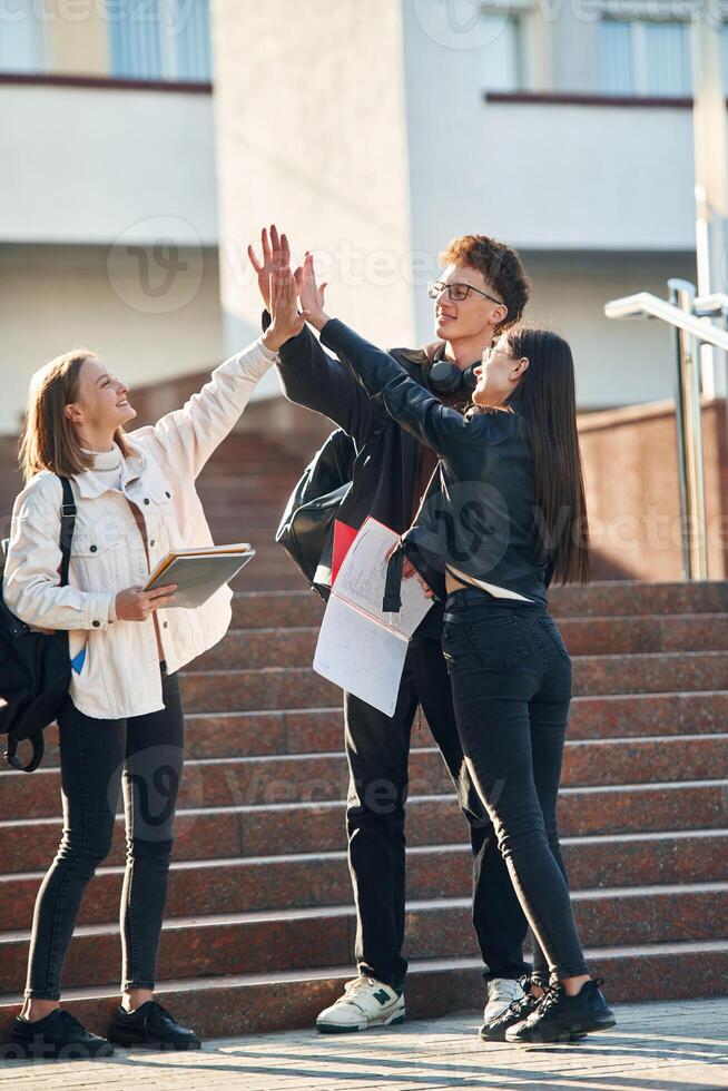 Giving high five to each other. Three young students are outside the university outdoors photo