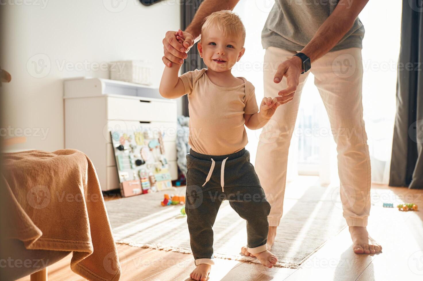 Boy is ,,walking by the help of dad. Man is playing with his little son in the domestic room photo