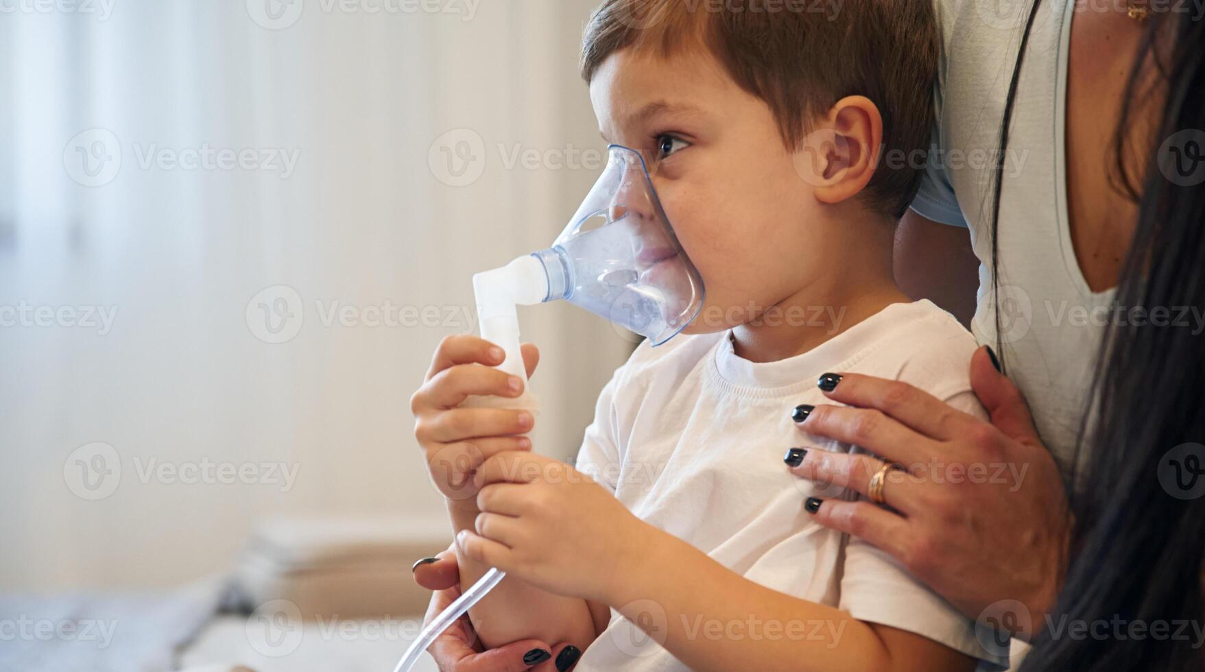 Boy is ,making, inhalation with nebulizer to reduce coughing. Mother with her little son is at home in domestic room photo