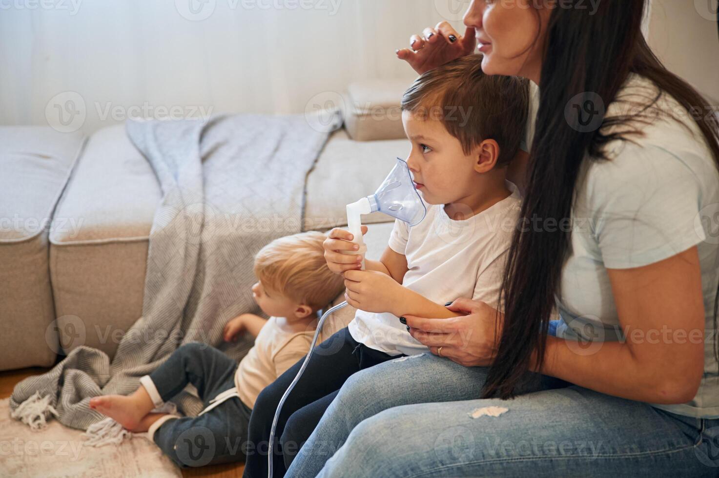 Boy is making inhalation with nebulizer, to reduce coughing. Mother with her little son is at home in domestic room photo