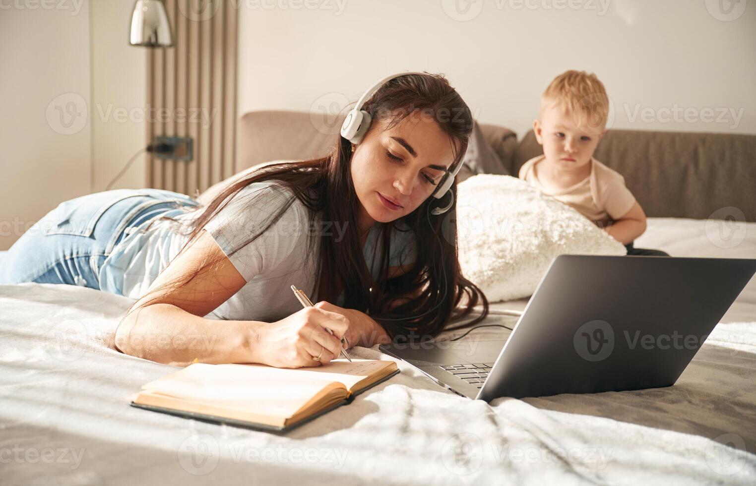 Woman ,in, headphones with laptop, writing in notepad. Mother with her little son is at home in domestic room photo