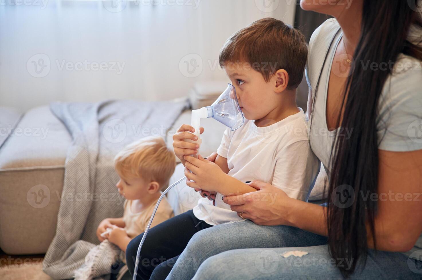 Boy, is making, inhalation with nebulizer to reduce coughing. Mother with her little son is at home in domestic room photo