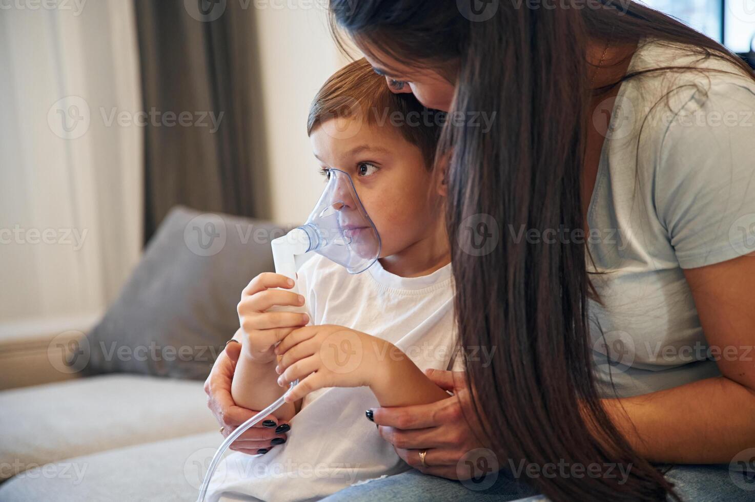 Boy is making, inhalation with nebulizer to reduce coughing. Mother with her little son is at home in domestic room photo