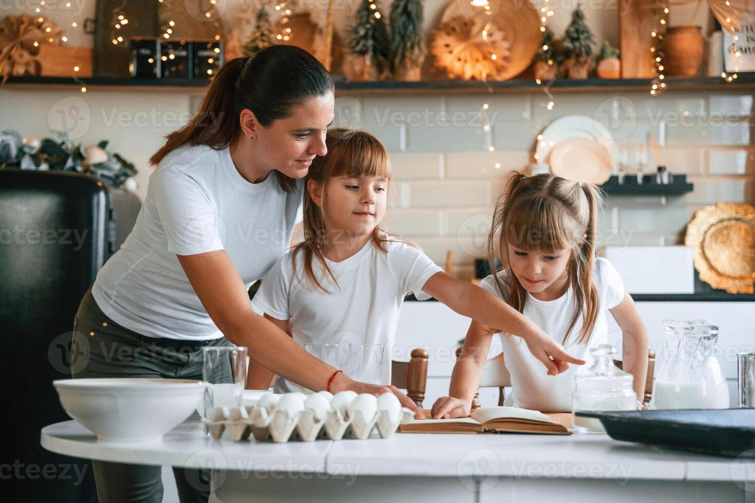 Learning how to prepare food... Mother with her little two daughters is on the kitchen with garlands photo