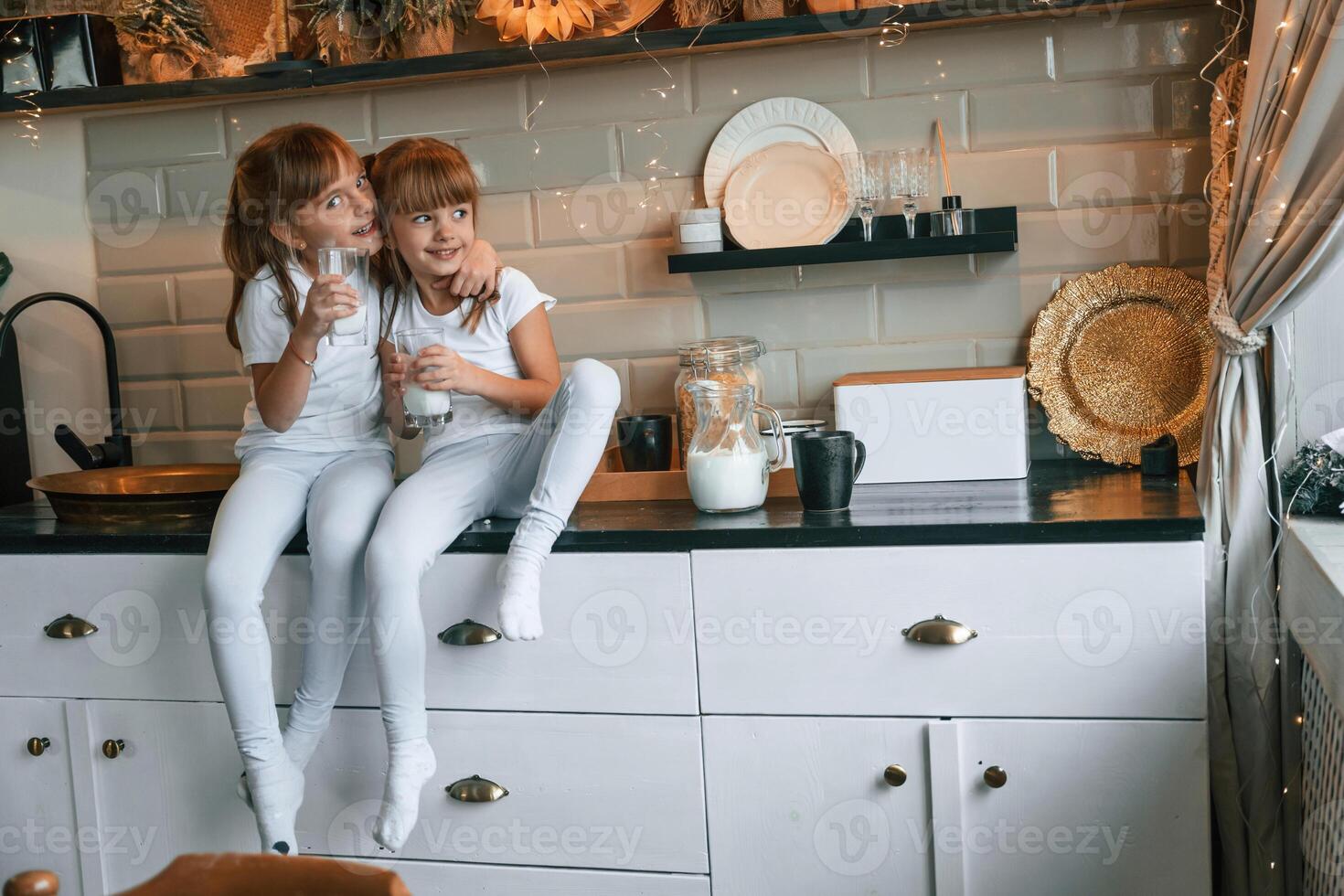 Happy two little ,,girls are drinking milk while sitting on the table on the kitchen photo