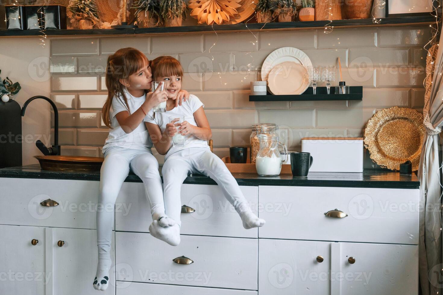 Happy two, little girls are drinking milk while sitting on the table on the kitchen photo
