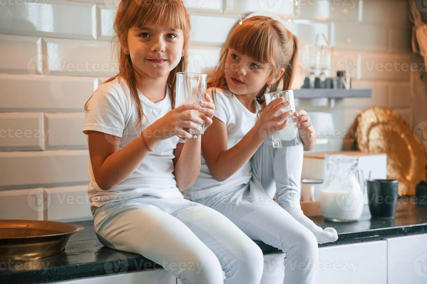 Having fun. ..Happy two little girls are drinking milk while sitting on the table on the kitchen photo
