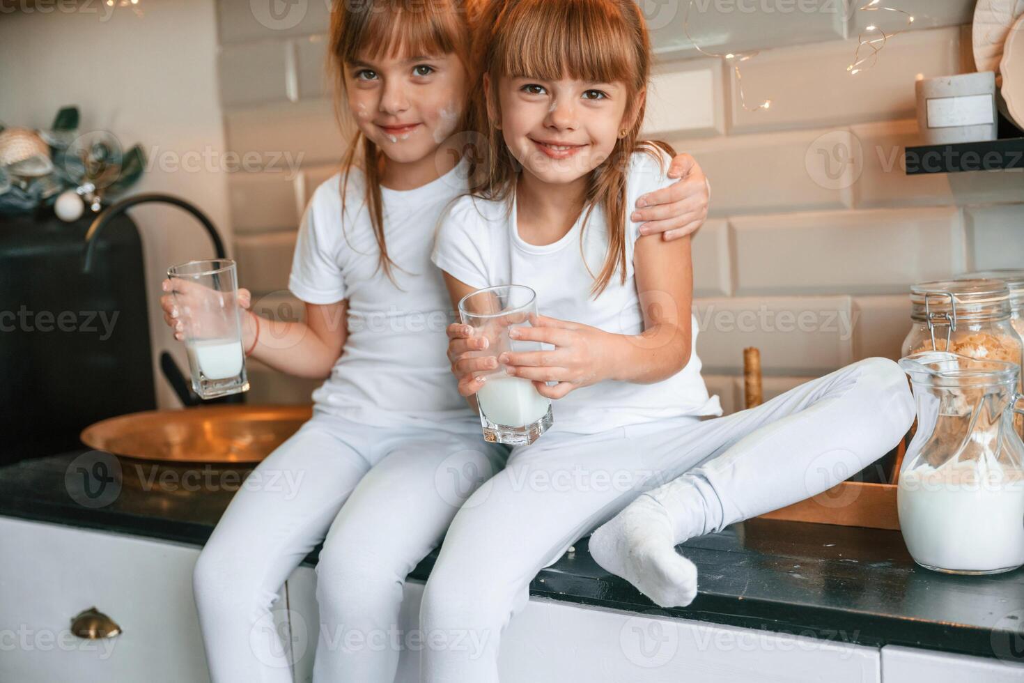 Embracing, each ,other. Happy two little girls are drinking milk while sitting on the table on the kitchen photo