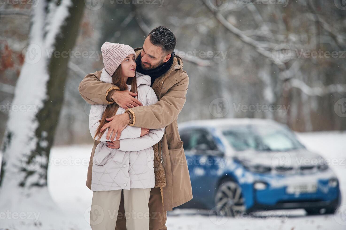 Standing ,and embracing.. Happy couple having a walk in winter forest. Blue car is parked photo