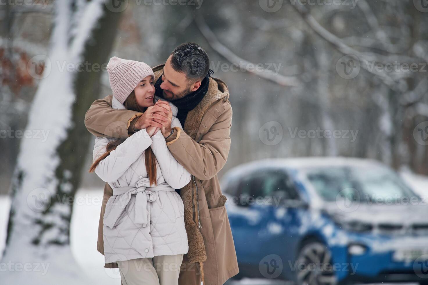 Standing and embracing... Happy couple having a walk in winter forest. Blue car is parked photo