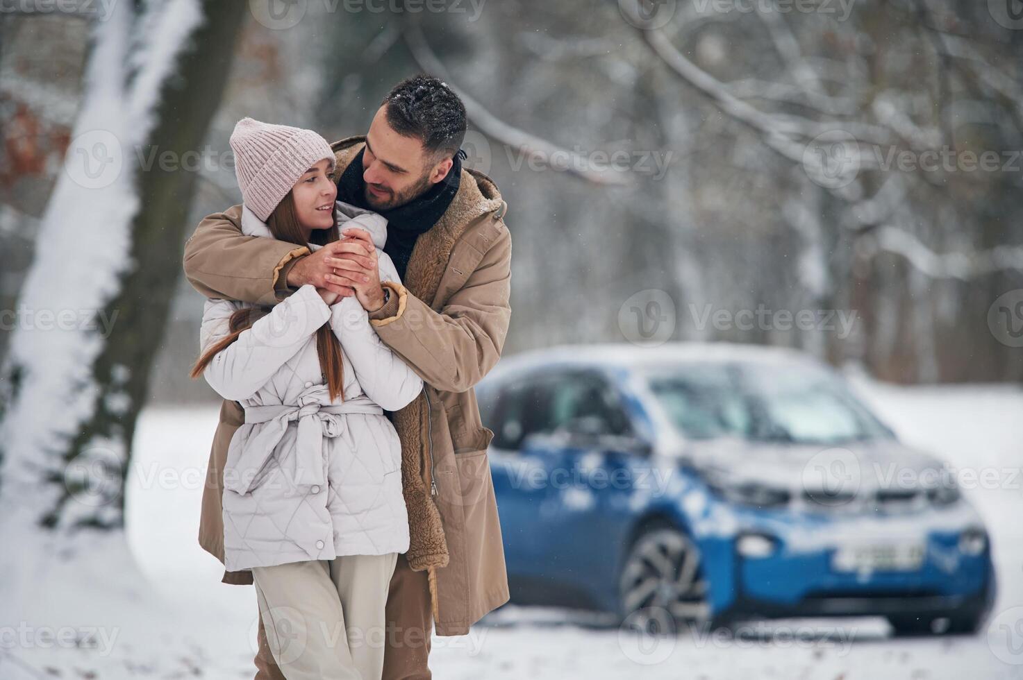 Standing and embracing. Happy, couple ,having a walk in winter forest. Blue car is parked photo