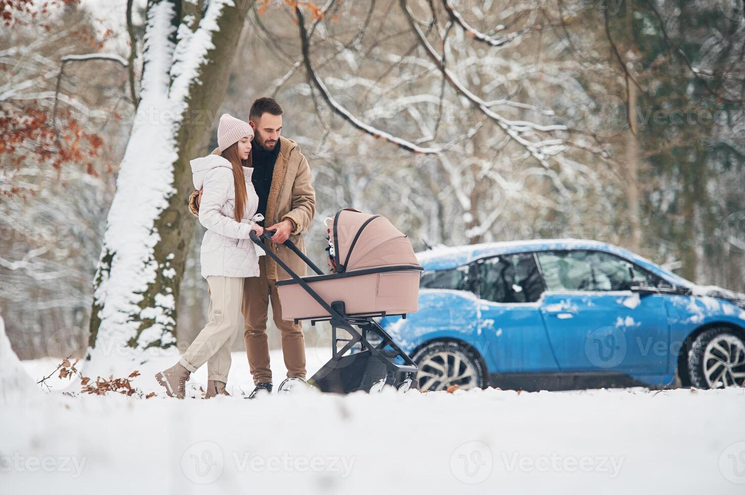 With ,,pram. Happy couple having a walk in winter forest. Blue car is parked photo