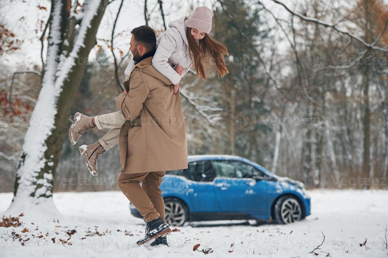 Playful mood.. Happy couple having a walk in winter forest. Blue car is parked photo