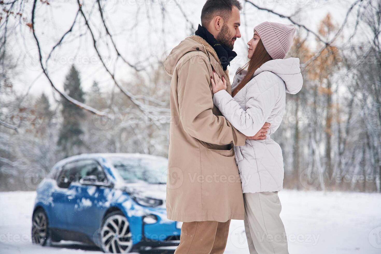 Standing and, embracing each other. Happy, couple having a walk in winter forest. Blue car is parked photo