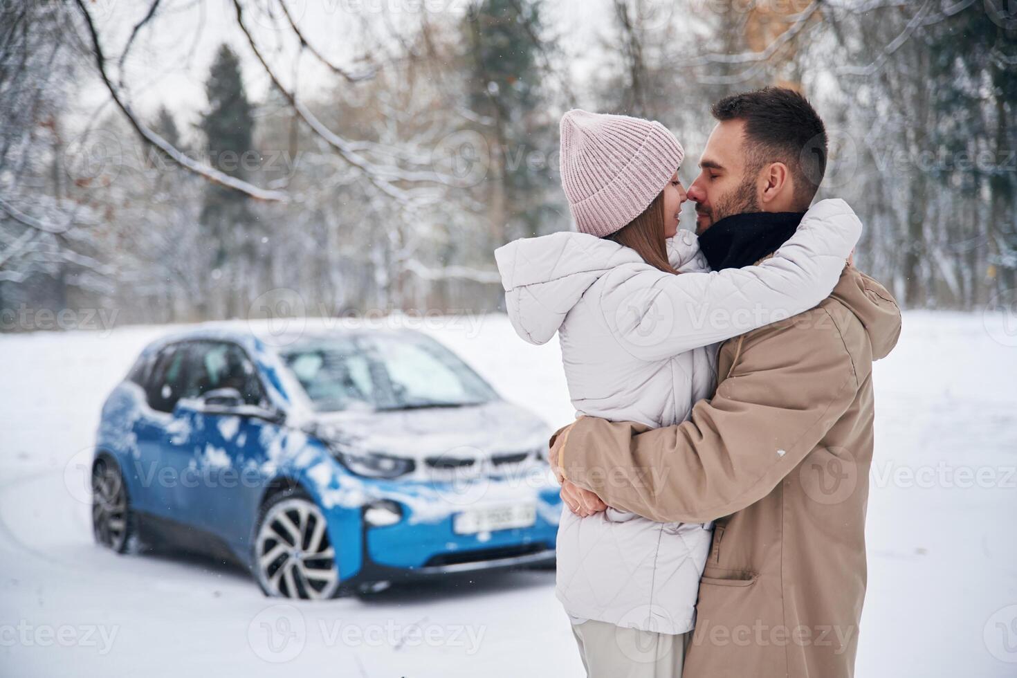 Standing, and, embracing, each other. Happy couple having a walk in winter forest. Blue car is parked photo