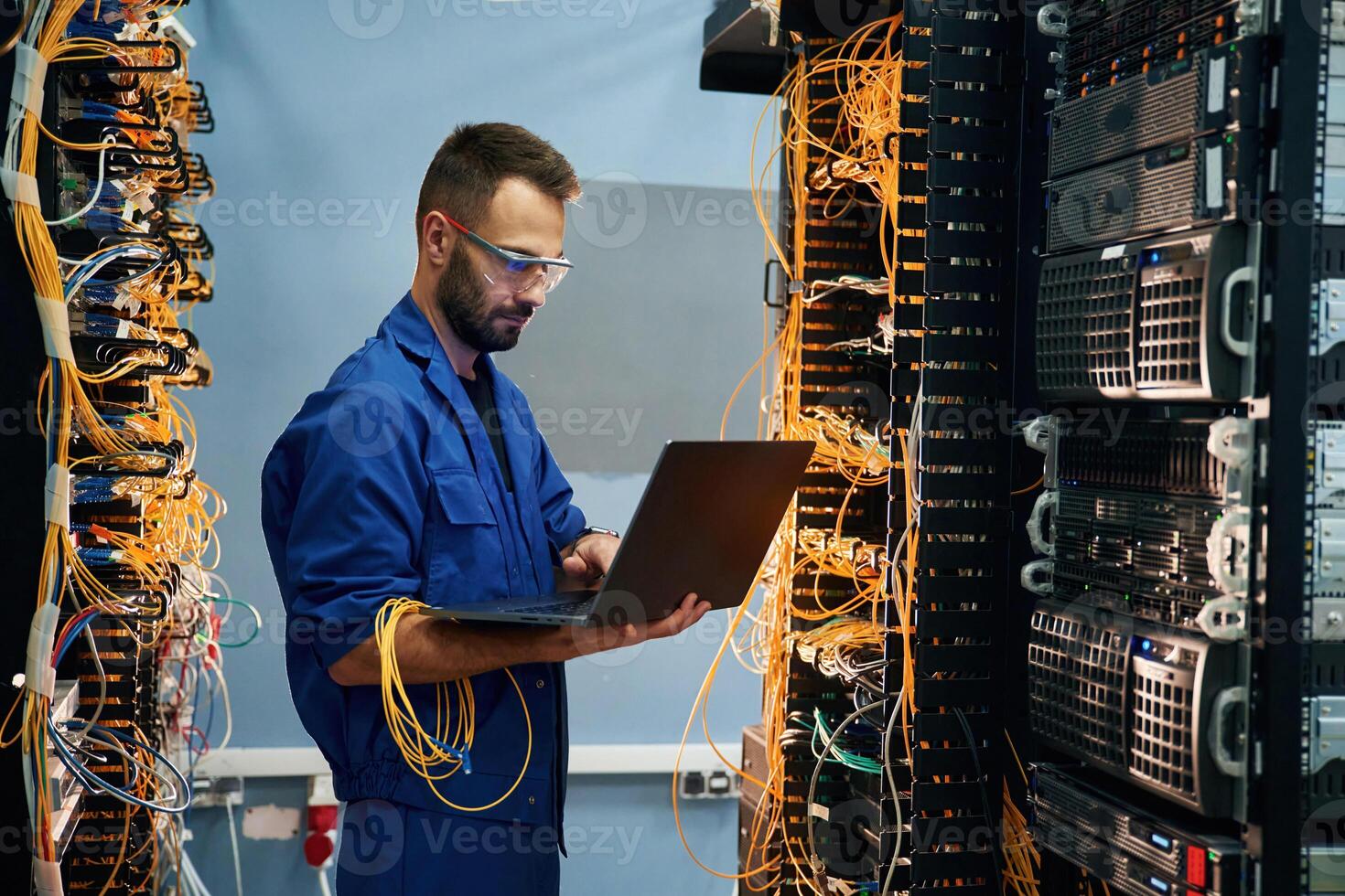 With laptop in hands,. Young man is working with internet equipment and wires in server room photo