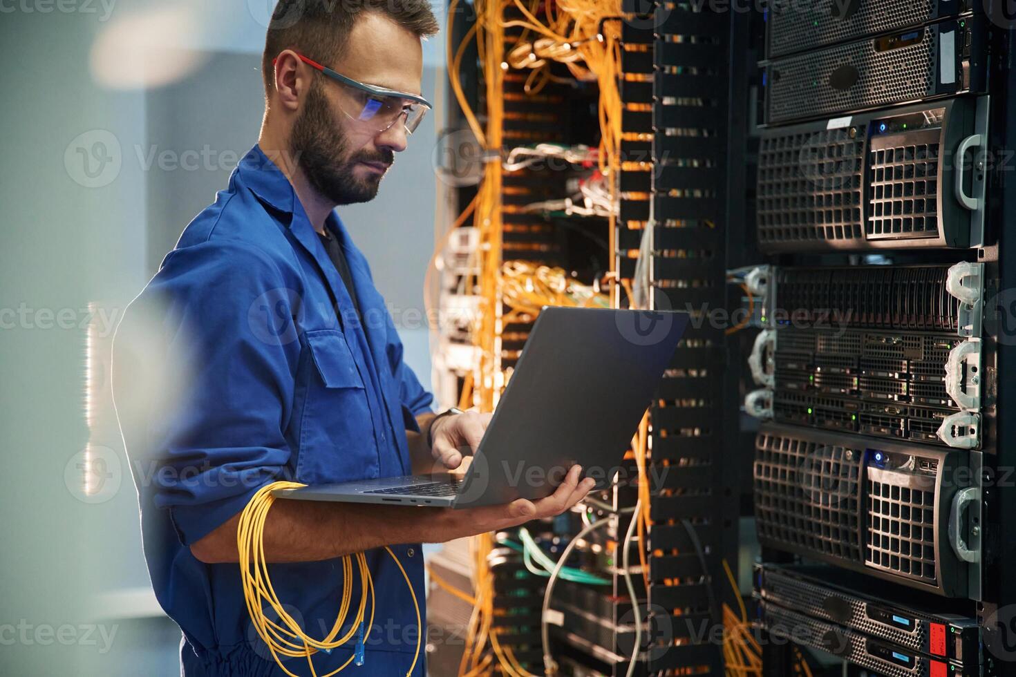Silver colored laptop in hands. Young man is working with internet equipment and wires in server room photo