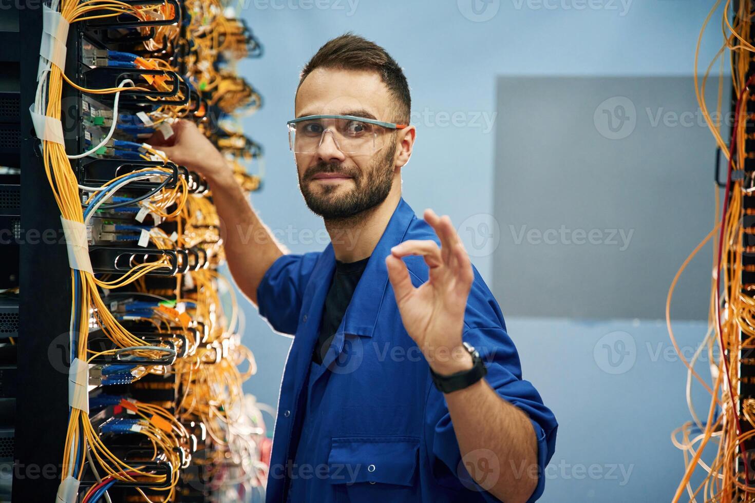 Okay or fine hand gesture. Young man is working with internet equipment and wires in server room photo