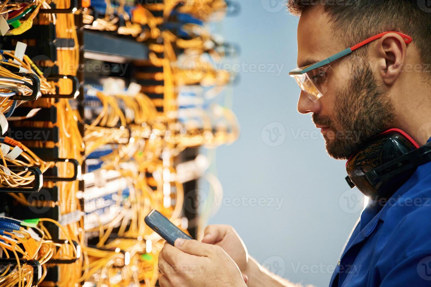 Holding ,smartphone.. Young man is working with internet equipment and wires in server room photo