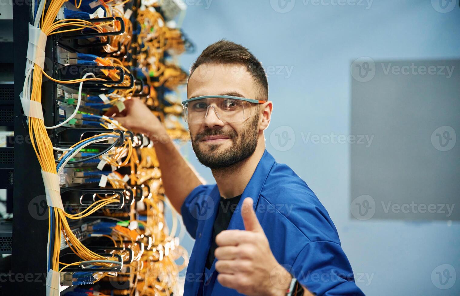 Fixed, with thumbs up.,. Young man is working with internet equipment and wires in server room photo