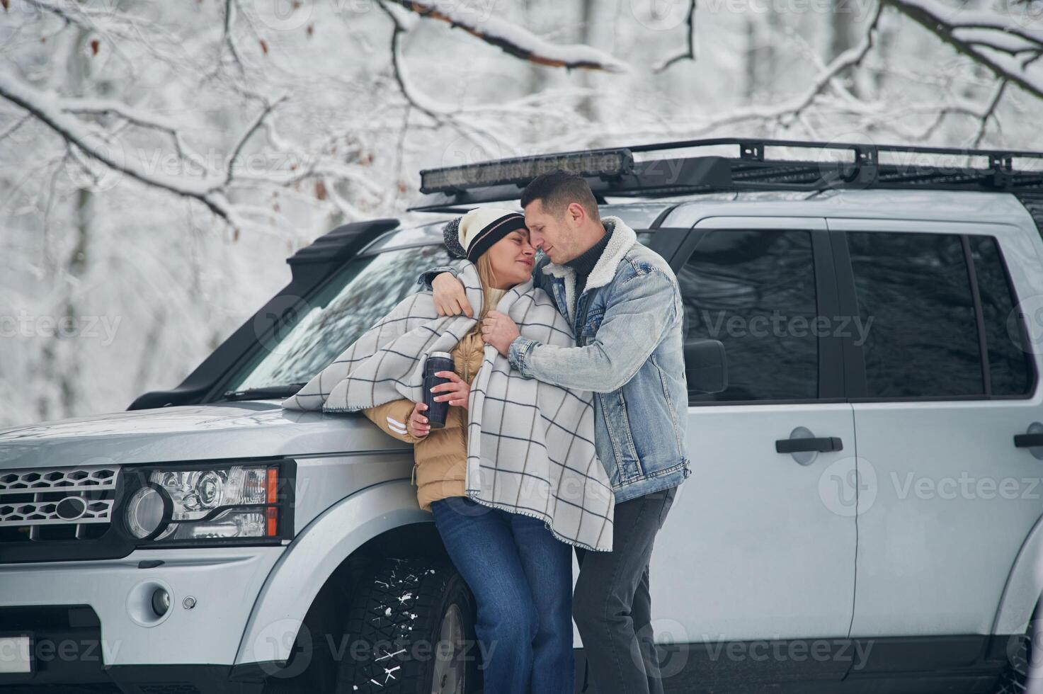 Man is covering woman with warm ,blanket.. Beautiful couple standing near the car in the winter forest photo