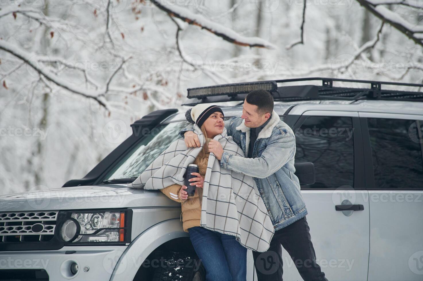 Man is covering ,woman with, warm blanket. Beautiful couple standing near the car in the winter forest photo