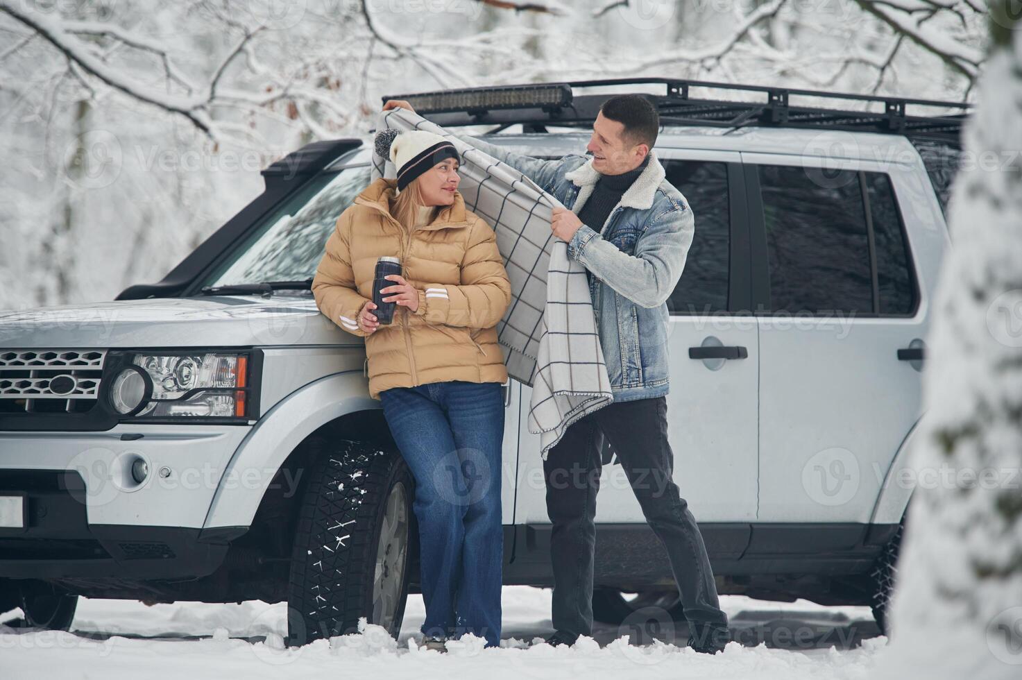 Man is, covering woman with warm blanket. Beautiful couple standing near the car in the winter forest photo