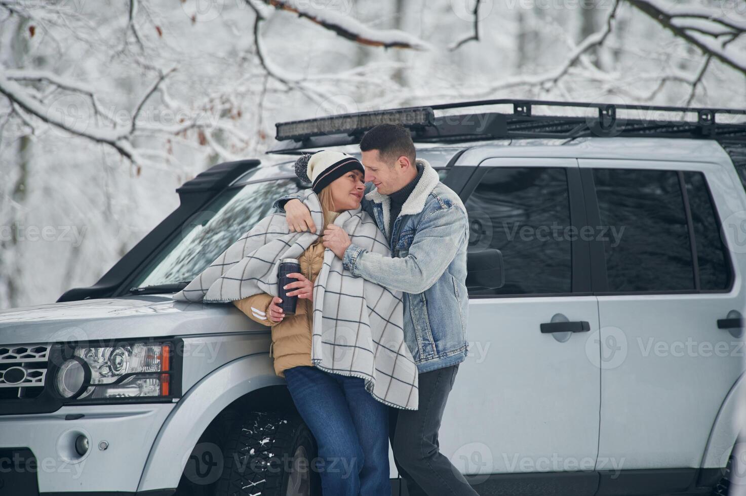 Man, is ,covering woman with warm blanket. Beautiful couple standing near the car in the winter forest photo