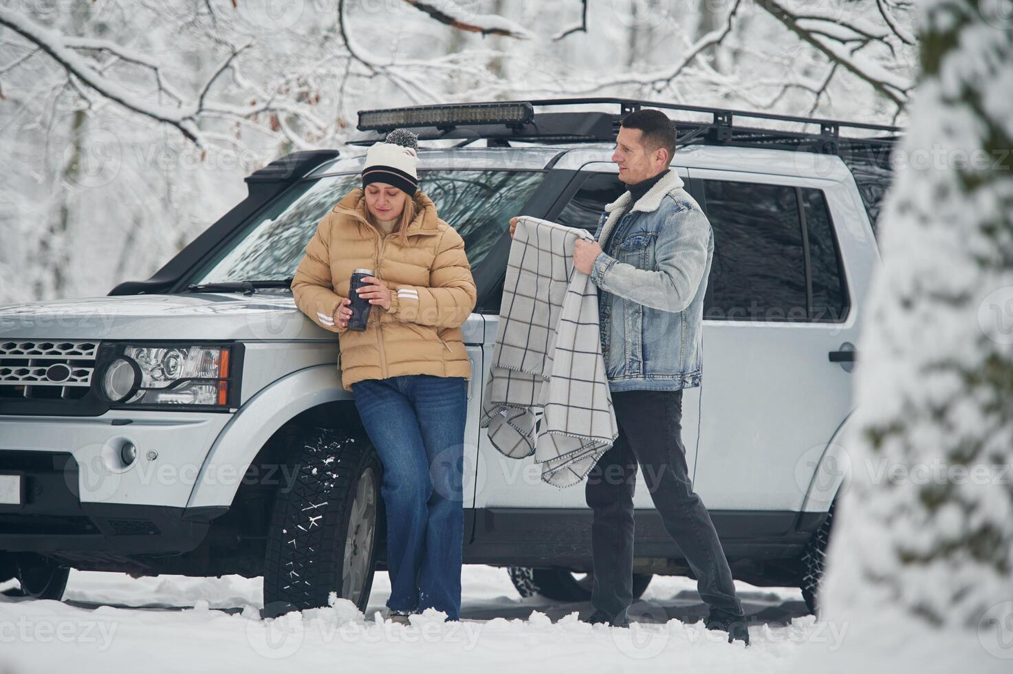 Man, is covering woman with warm blanket. Beautiful couple standing near the car in the winter forest photo