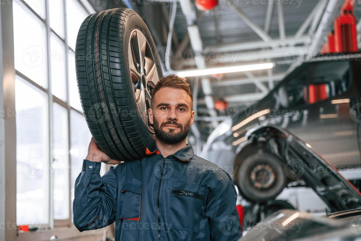 Holding, a, tire. Car repairman is in the garage with automobile photo
