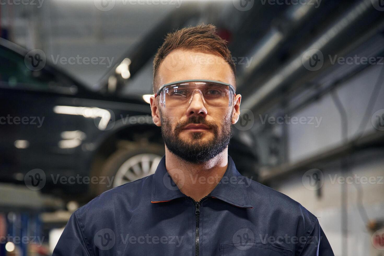 Looking, at the camera. Car repairman is in the garage with automobile photo