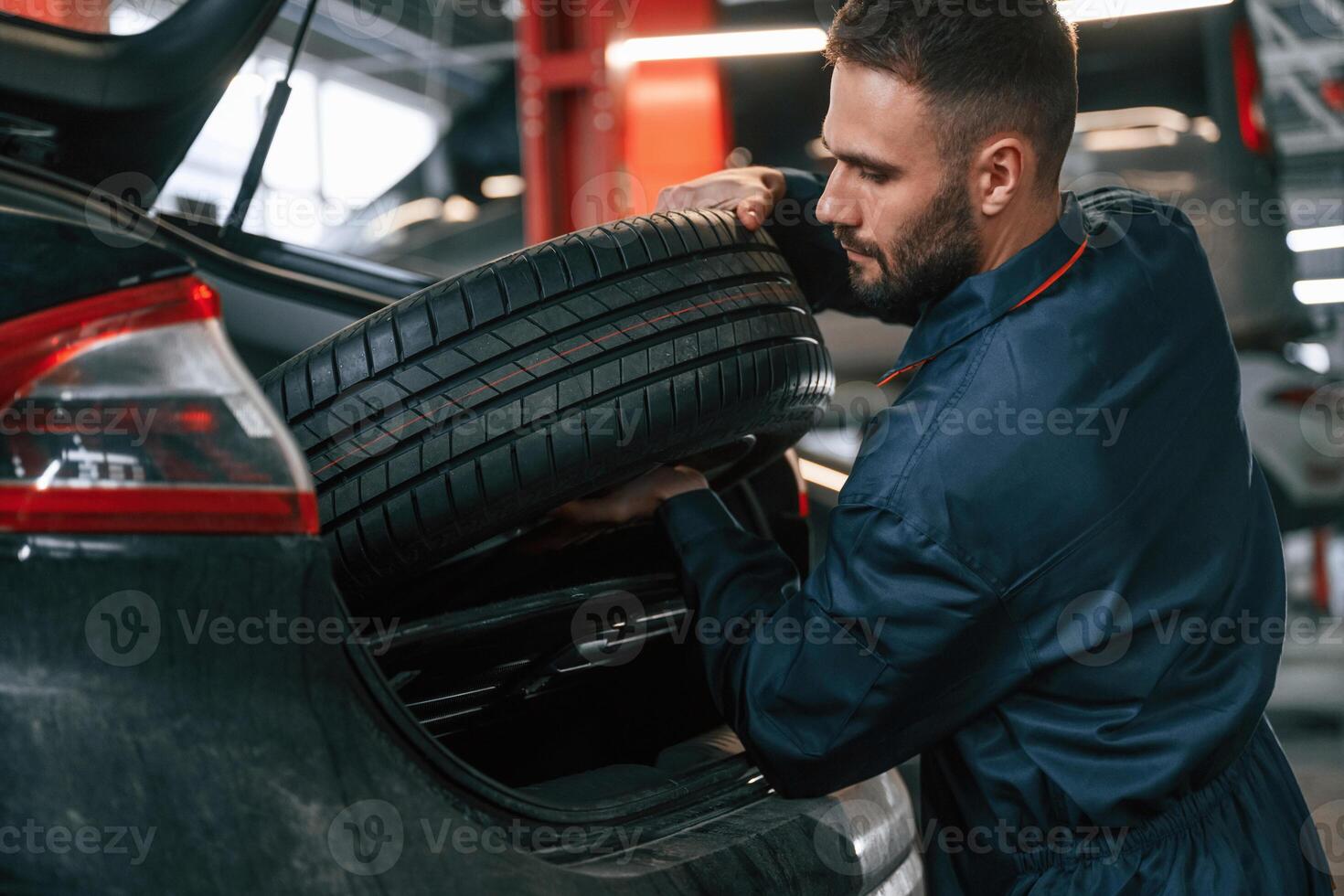 Holding ,a tire. Car repairman is in the garage with automobile photo