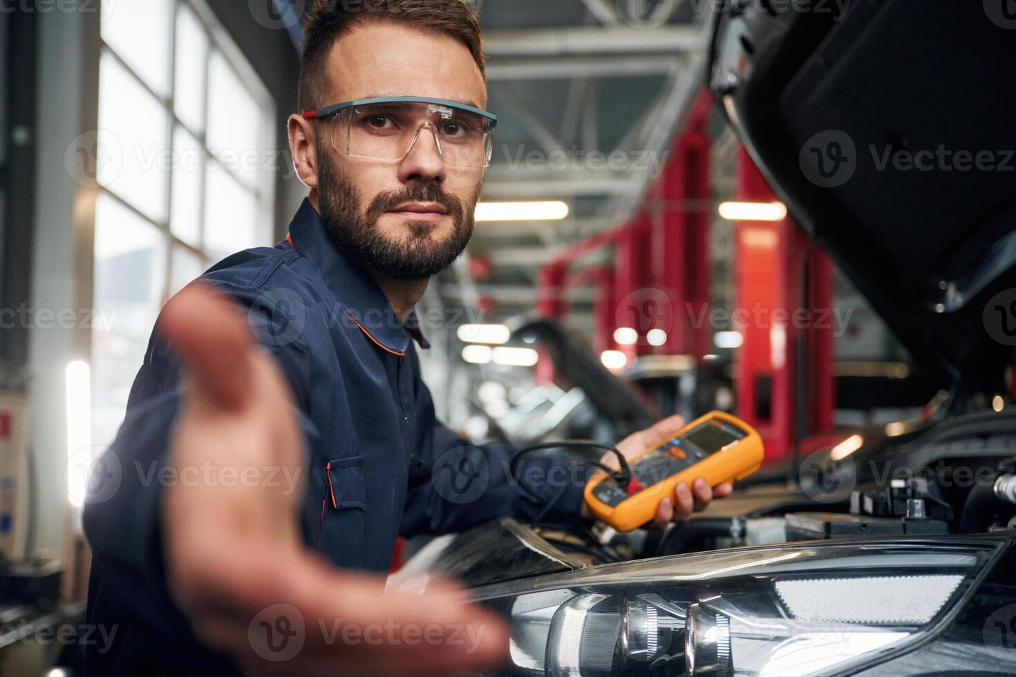 Yellow, colored device for measuring electricity is in hands. Car repairman is in the garage with automobile photo
