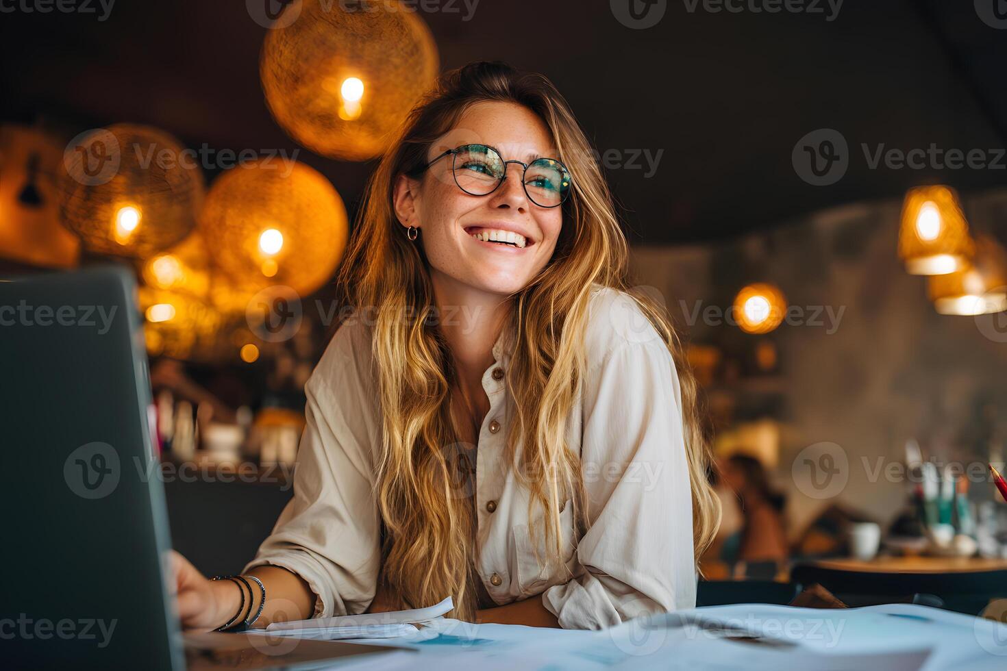 Young woman wearing stylish glasses sits at a cafe table, smiling as she focuses on her work. photo