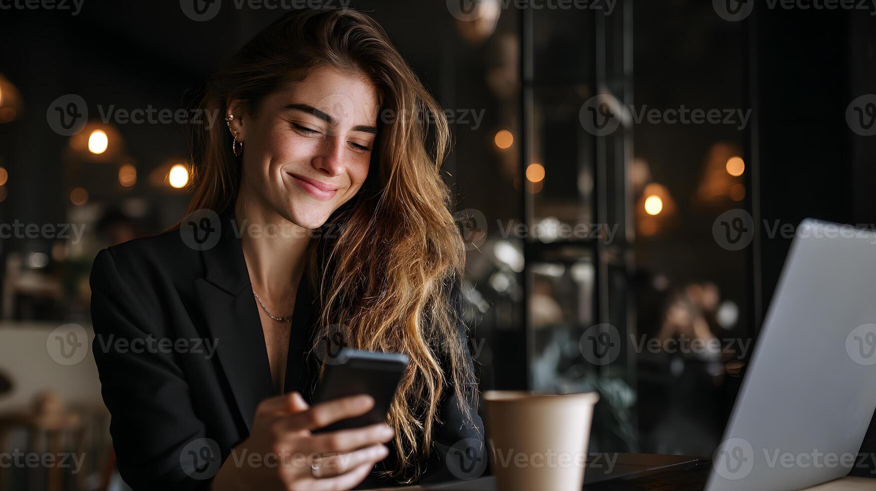 Smiling young woman in a stylish black blazer engaging with her smartphone while sitting at a table. photo