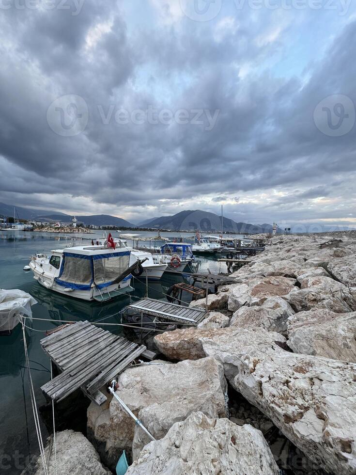 Boats anchored at harbor with rocky shoreline under cloudy sky in evening light photo
