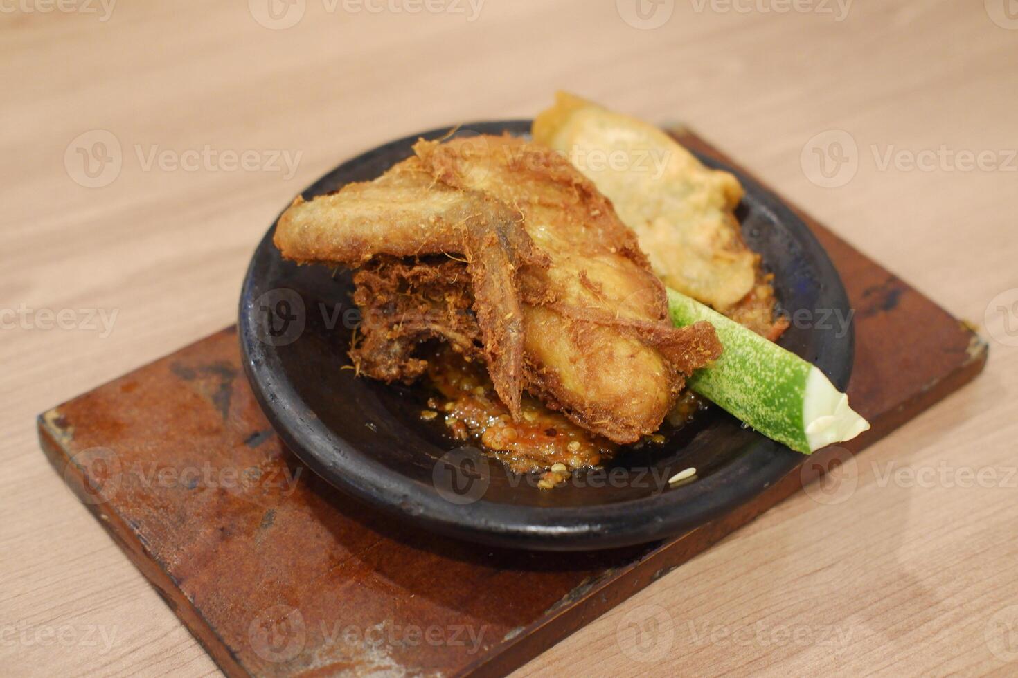 The dish features a piece of crispy fried chicken served on a traditional stone plate. It sits atop a spicy sambal chili sauce, adding a vibrant and flavorful base photo