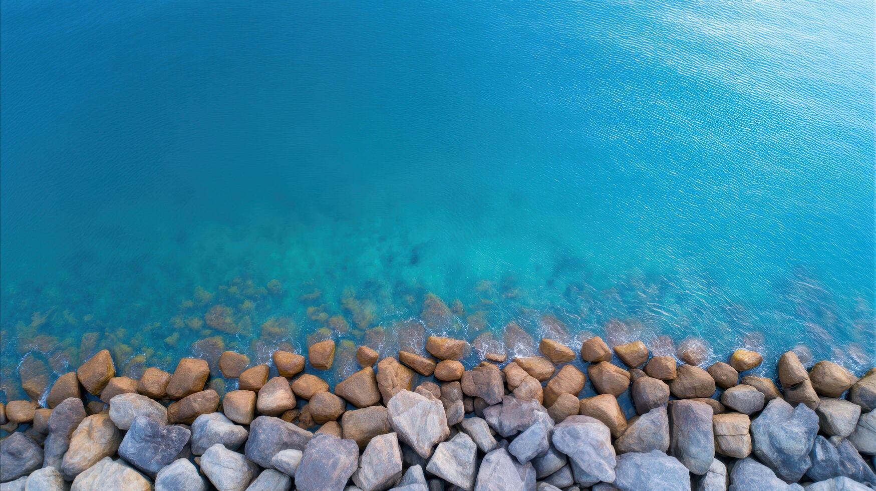 Aerial view of turquoise water with rocky shoreline and visible underwater stones along a calm coastal edge under sunlight. photo