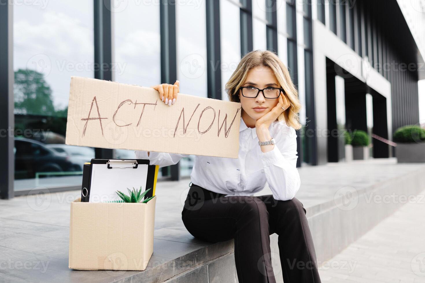 Woman sitting outdoor with poster Act Now next to box of stuff . Beautiful female protester at strike against unemployment photo