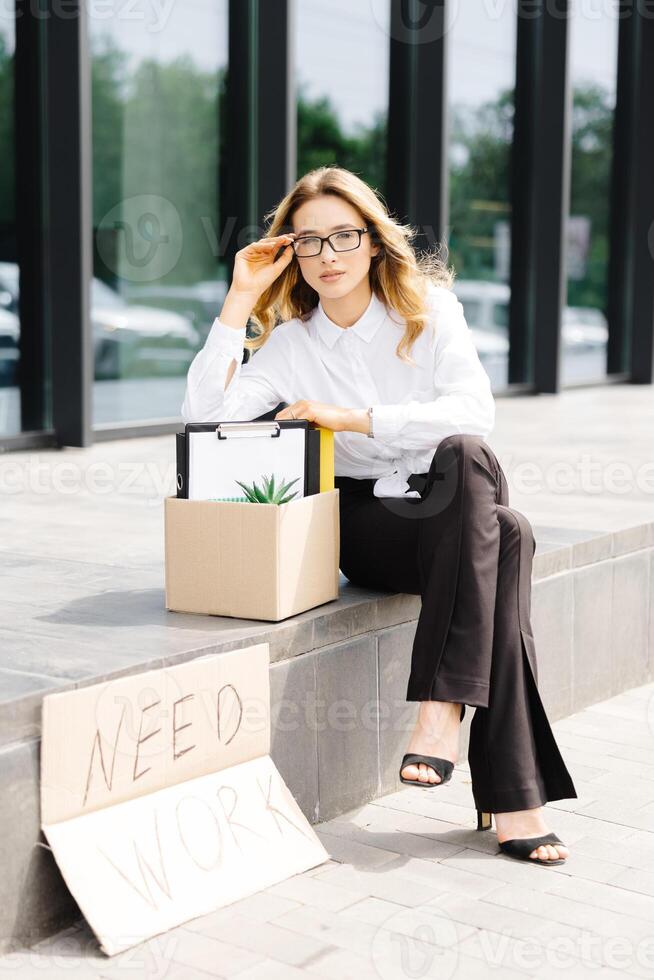 Woman sitting with cardboard banner with the slogan Need work next to box of stuff. Beautiful female protester at strike against unemployment photo
