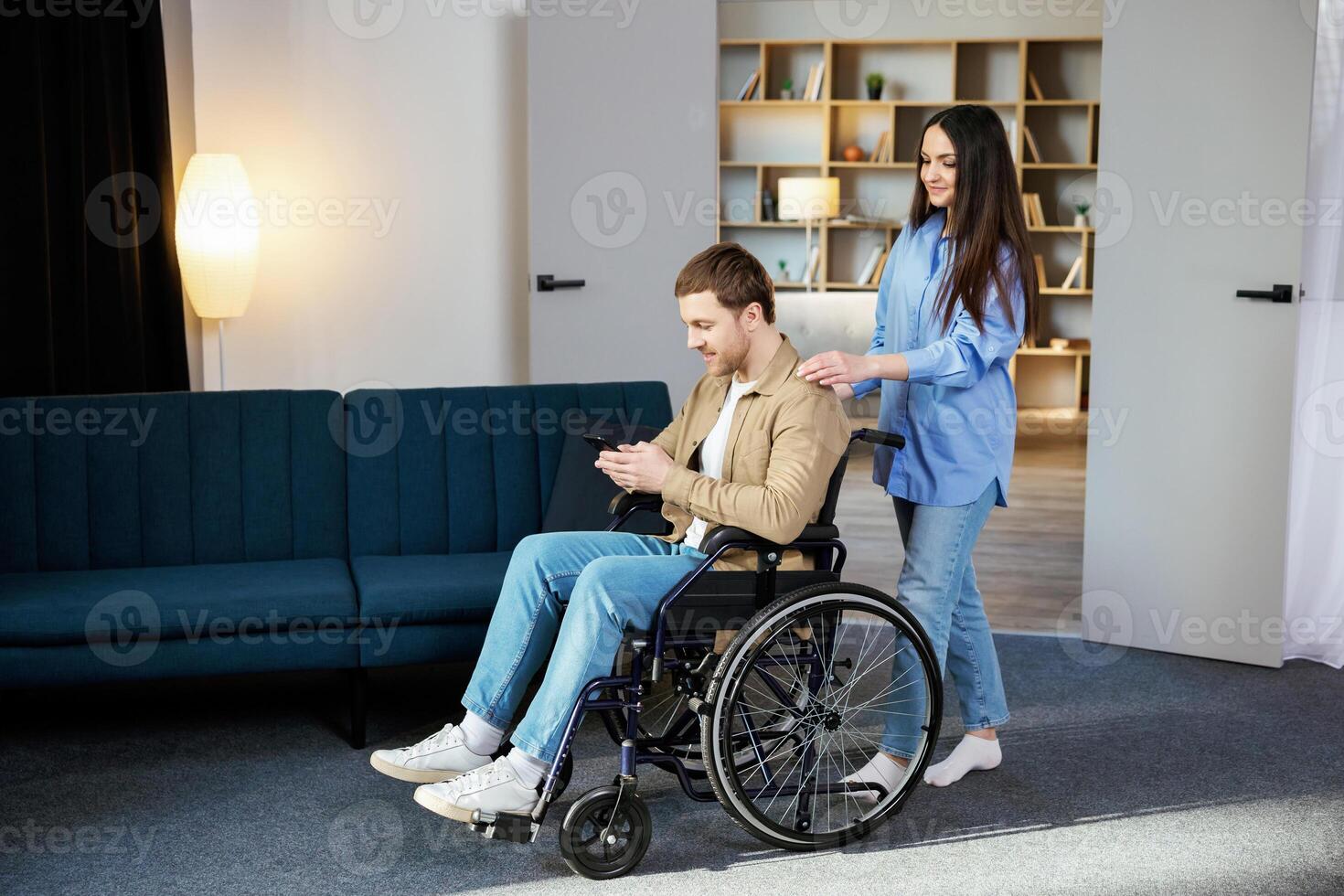 Young man in a wheelchair is holding a mobile phone while his girlfriend helps him move around the apartment photo