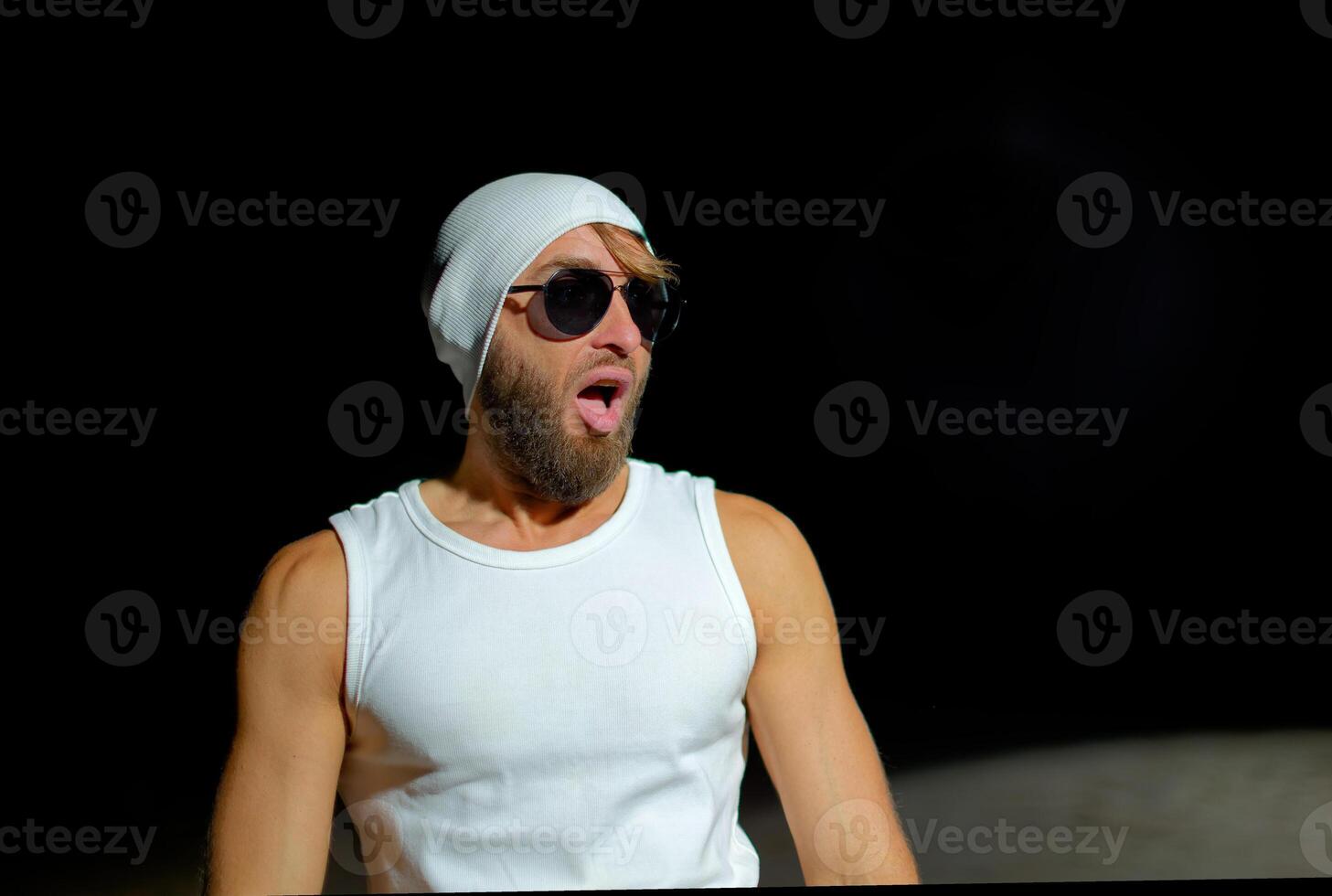 Man wearing a tank top, sunglasses, and a beanie poses confidently at night against a dark background. photo